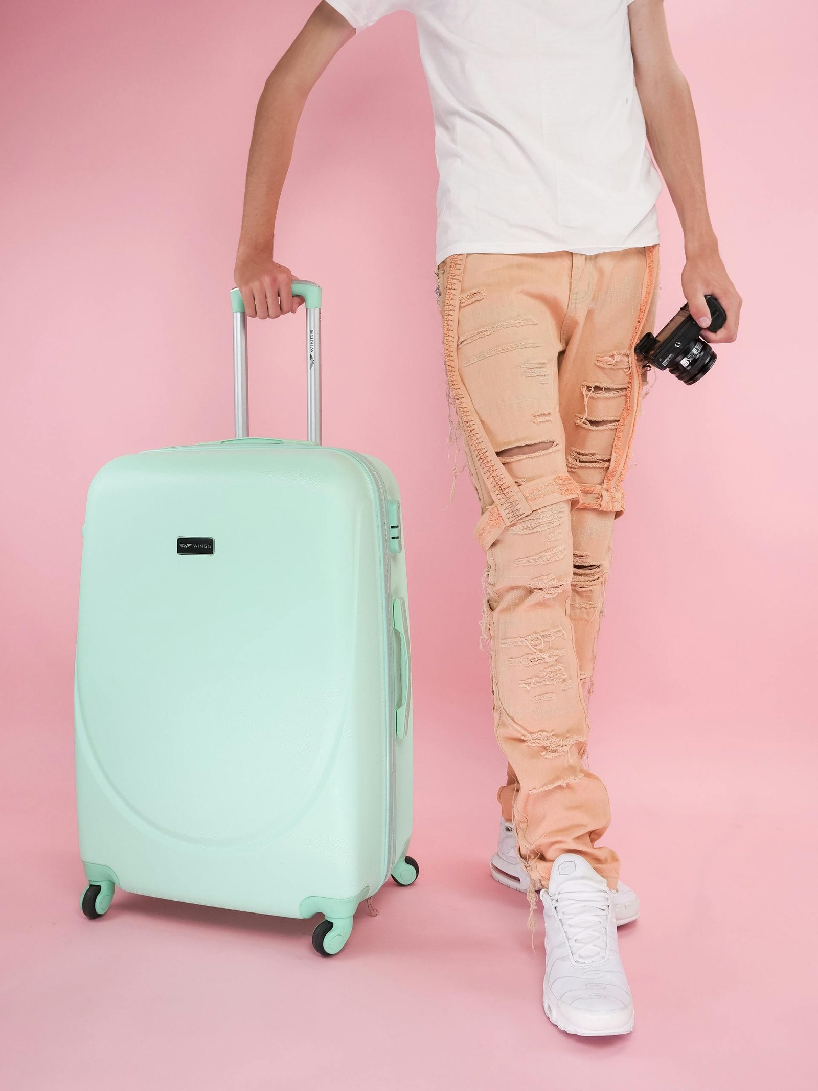 Adult holding a mint suitcase and camera, ready for travel against a pink backdrop.